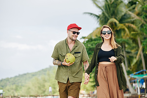 Happy young Caucasian couple in sunglasses holding hands when walking on the beach in tropical country