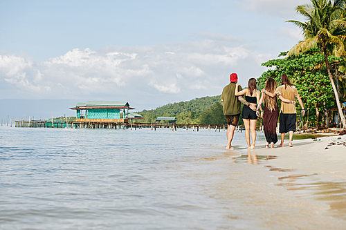 Group of young hugging people walking along seashore in tropical country, view from the back