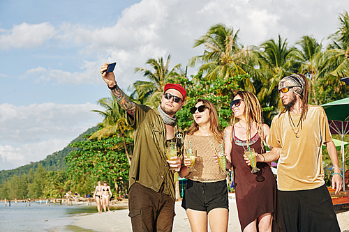 Group of joyful young people walking on beach, drinking sweet cocktails and talking selfie together