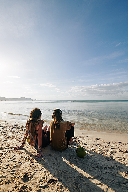 Young couple sitting on sandy beach, drinking coconut water and looking at beautiful calm sea