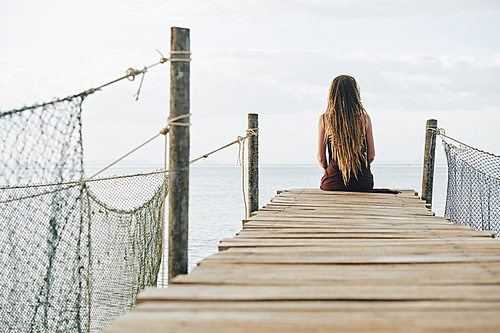 Pensive young woman with dreadlock sitting on narrow handmade wooden pier and looking at beautiful calm ocean