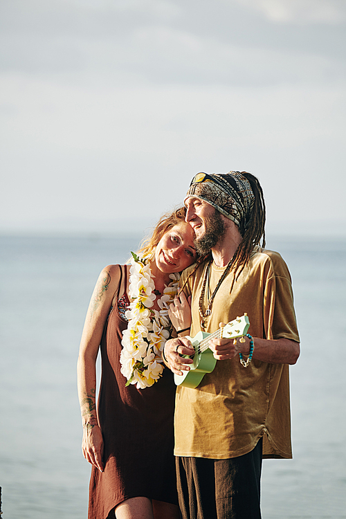 Positive man with dreadlocks playing ukulele and singing a song for his happy beautiful wife when they are spending time on the beach