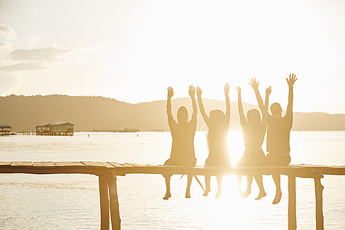 Group of happy excited friends sitting on handmade wooden pier, shouting and raising arms in sunset sun rays, backlit
