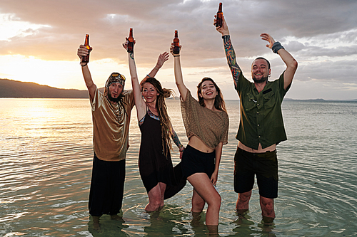 Happy friends dancing in water and raising hands with beer bottles when having fun at beach party