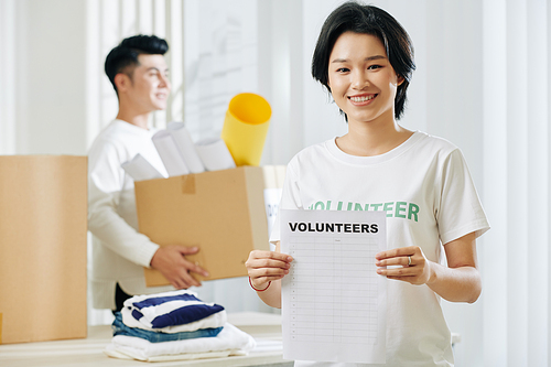 Portrait of pretty young Asian woman standing in donation center with list of volunteers in hands