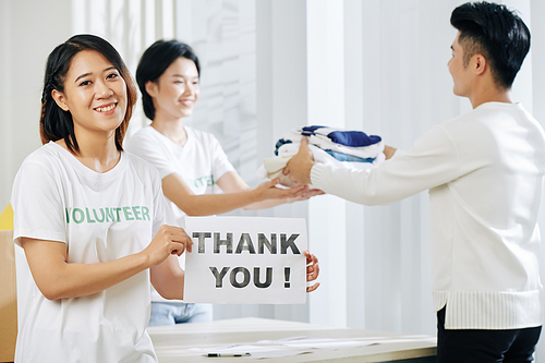 Positive young pretty Asian volunteer working at donation center and showing thank you card