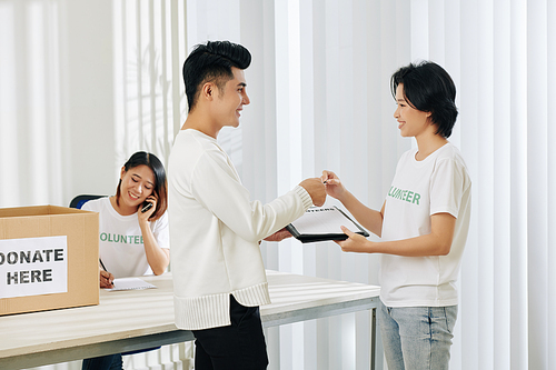 Volunteer asking smiling young Asian man to sign up for volunteer work in donation center