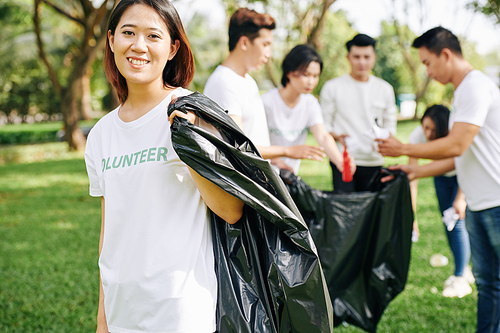 Portrait of young happy volunteer with big plastic trash bag for collecting garbage in park