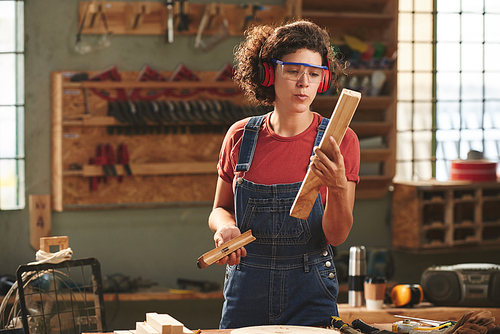 Professional carpenter at work. Young curly woman in protective eyewear, earmuff and denim overall checking quality of processed wooden plank