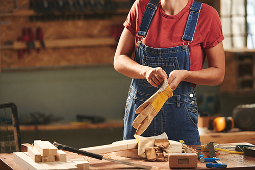 At manufacturing workshop. Unrecognizable female carpenter in denim overall putting on gloves before polishing wooden planks