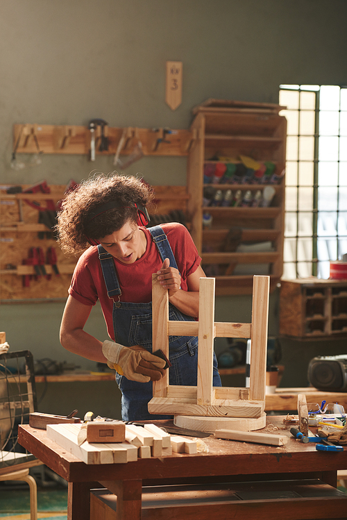 Woodworking process. Concentrated female carpenter in earmuffs and protective gloves carefully polishing wooden stool lying upturned on table