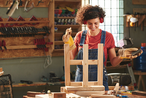 Carpenter in workshop. Young curly woman in denim overall, earmuffs and protective gloves looking at finished wooden stool after polishing and smiling brightly