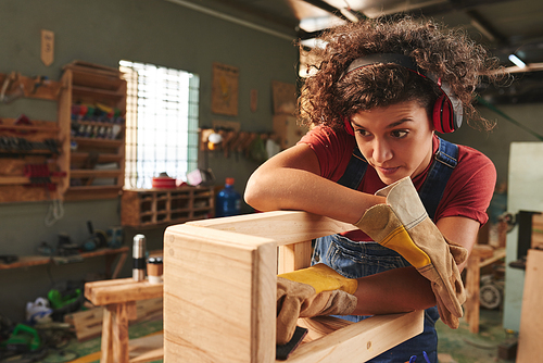 At carpenter workshop. Young curly woman in ear defenders and protective gloves checking quality of wooden stool after polishing it