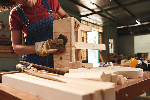 Woodworking process. Young professional female carpenter in denim overall and protective gloves carefully polishing wooden stool with sandpaper, low angle view