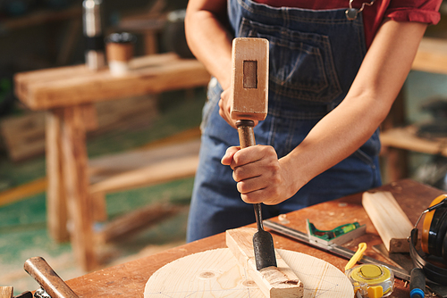 At manufacturing workshop. Close-up view of female carpenter striking chisel with hammer into wooden plank for removing wood pieces