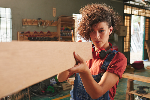 Professional carpenter at work. Young concentrated woman with curly hair holding wooden plank and measuring its length with her eyes