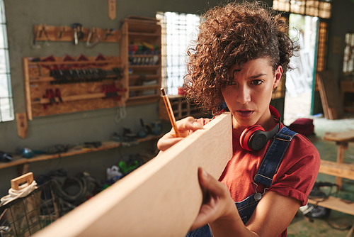 Measure twice and cut once. Young concentrated female carpenter with curly hair holding wooden plank and estimating its length before sawing