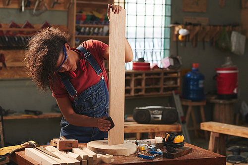 At carpenter workshop. Young curly woman in denim overall and safety glasses enjoying process of polishing wooden plank with sandpaper