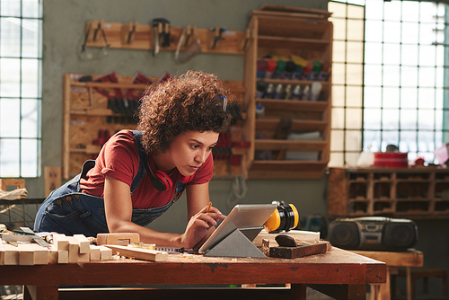 Carpentry for beginners. Young concentrated woman with curly hair reading instructions on digital tablet before working with wood