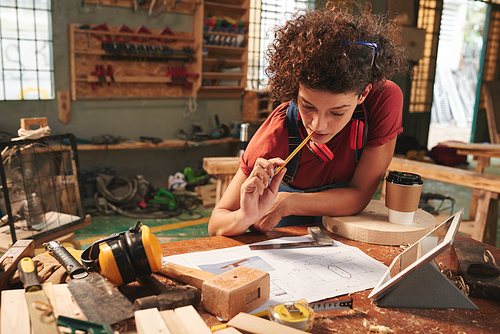 Professional carpenter at work. Young curly woman leaning over messy table and looking at draft with new project thoughtfully with pencil in her mouth