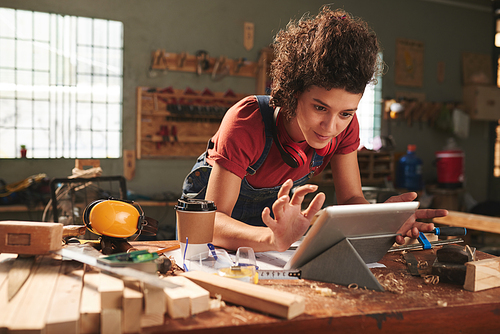 Carpentry for beginners. Young pretty woman with curly hair watching woodworking tutorial on digital tablet while leaning on messy table covered with sawdust