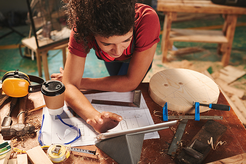 High angle view of young female carpenter reading manual on digital tablet while leaning over table covered with drafts, tool and sawdust