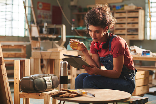 At carpentry workshop. Young pretty female carpenter in denim overall resting on wooden bench with cup of tea and reading book on digital tablet