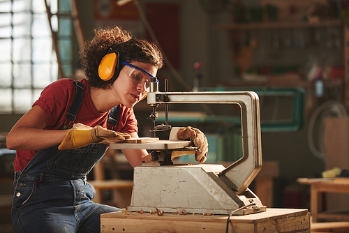 Professional carpenter at work. Young concentrated woman in protective eyewear and earmuffs making holes in wooden plank with drill press