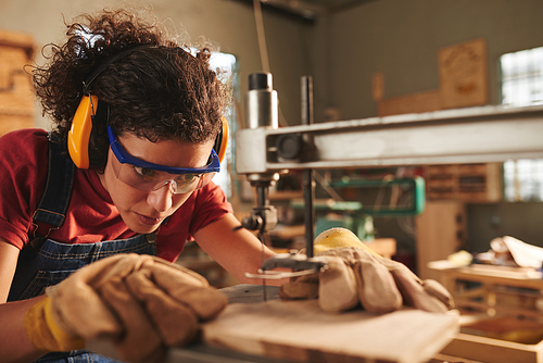 Woodworking process. Close-up view of young concentrated female carpenter in safety glasses and ear defenders drilling hole in wooden plank with drill press