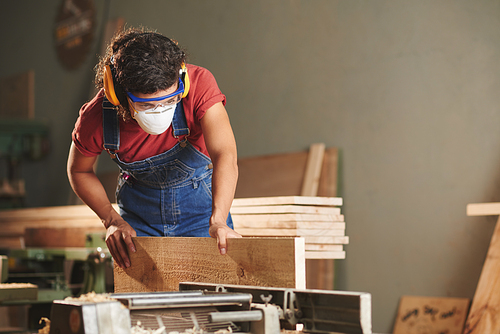Professional carpenter at work. Concentrated woman in denim overall, protective eyewear and earmuffs processing wooden plank on woodworking machine