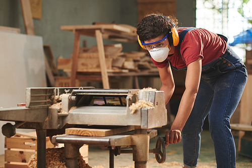 Woodworking process. Concentrated female carpenter in mask, safety glasses and earmuffs processing wooden board on woodworking machine