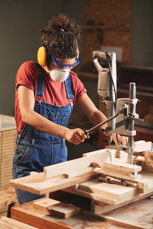 Woodworking industry. Young female carpenter in protective eyewear and ear defenders pressing lever on woodworking machine while cutting wooden planks