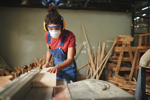 At manufacturing studio. Professional female carpenter in protective eyewear, earmuffs and mask cutting wooden board on table saw