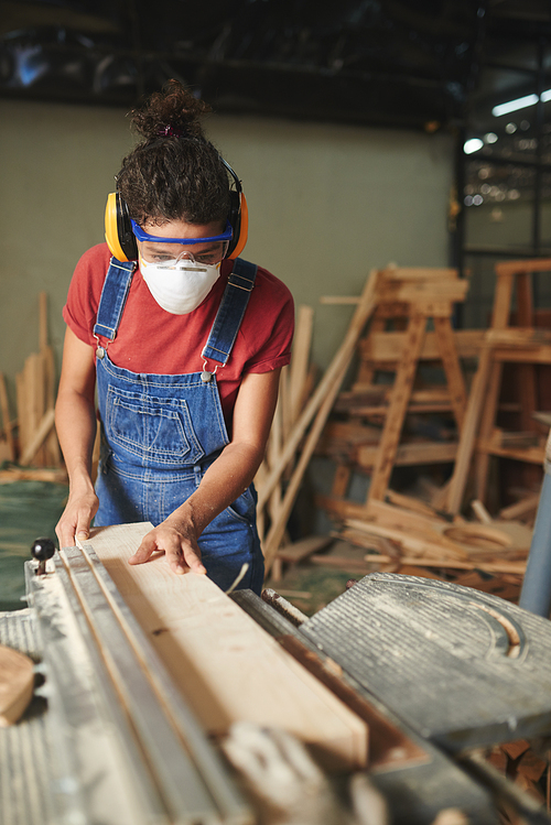 At carpentry workshop. Concentrated young woman in safety glasses, mask and earmuffs cutting wooden board on table saw