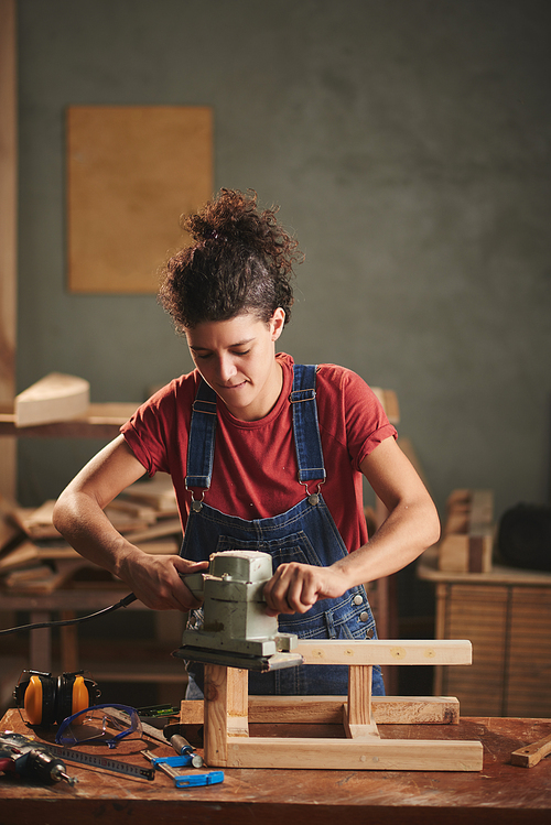 At carpentry workshop. Young curly woman in denim overall enjoying process of smoothing wooden stool surface with electrical belt sander