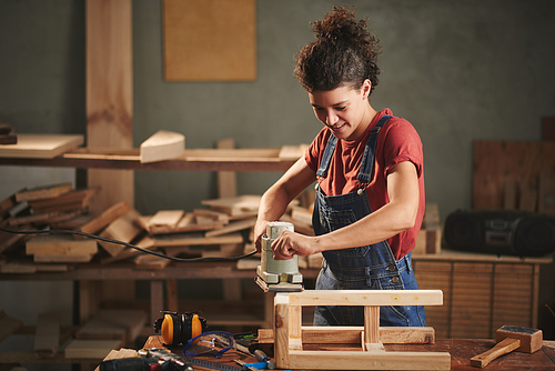 In love with woodworking. Young pretty female carpenter carefully smoothing wooden stool with electrical belt sander and smiling