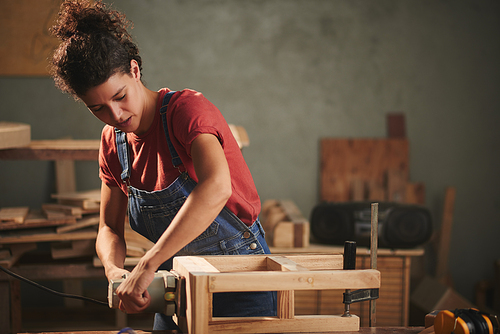 Professional carpenter at work. Young concentrated woman in denim overall smoothing just finished wooden stool with electrical belt sander