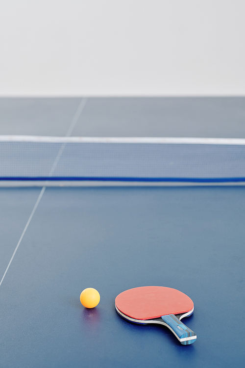 Ping pong racket and orange ball on table divided into two halves by a net, focus on foreground