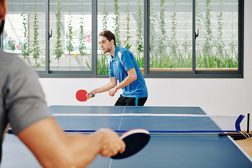 Handsome young sportsman playing ping pong with friend and hitting ball with racket
