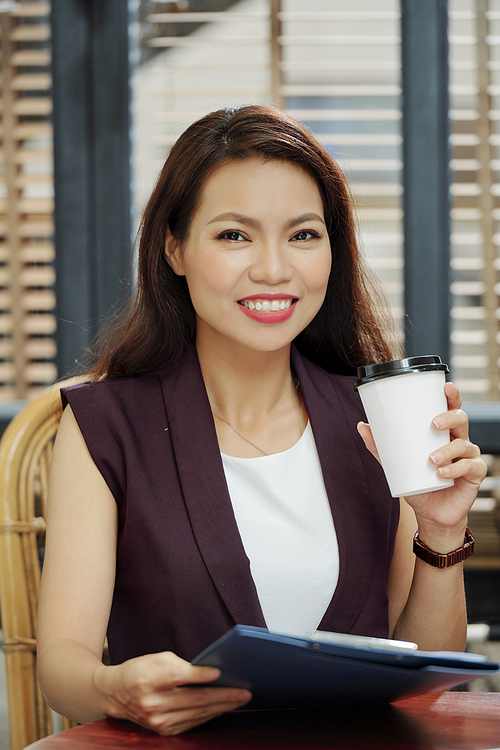 Attractive young Vietnamese businesswoman drinking cup of take away coffee and reading document on clipboard