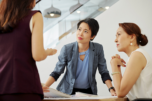 Frowning serious Asian female business executives looking at colleague telling her questionable ideas