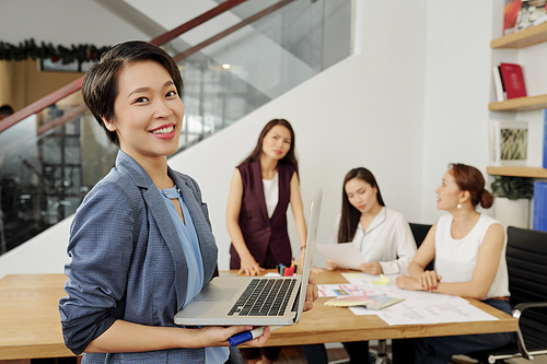 Pretty young Asian businesswoman standing with opened laptop and smiling at camera, her colleagues discussing project in background