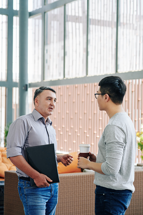 Serious mature entrepreneur with leather folder in hand talking to young employee during coffee break