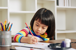 Serious little Vietnamese girl concentrated on drawing with red pencil when sitting at table at home