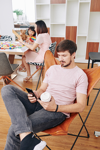 Young man drinking coffee and checking social media while his wife playing with daughter in background