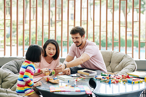 Cheerful multi-ethnic parents playing with little daughter, giving her puzzle toys and bricks
