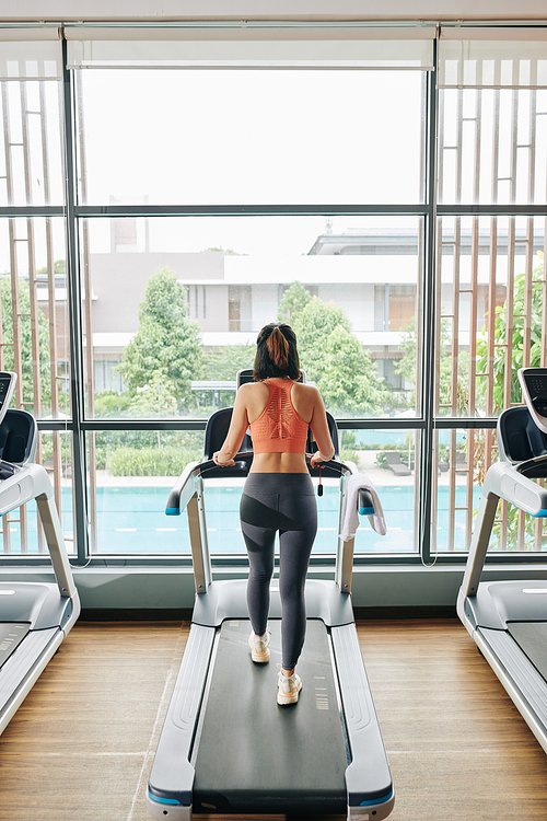 Slim young woman walking on treadmill in gym with big panoramic windows