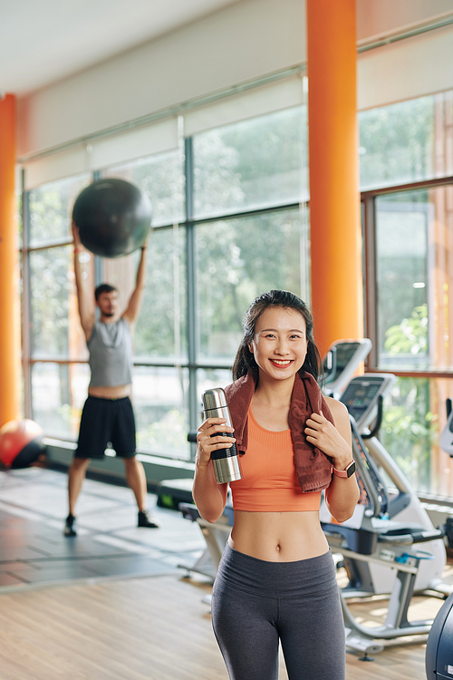 Beautiful happy young Vietnamese woman with towel and small thermos standing in gym after training in the morning