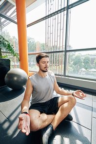 Calm young man sitting in lotus position on gym floor, keeping hands in mudra and meditating