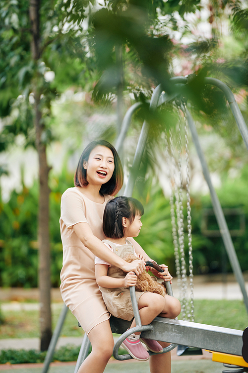 Cheerful young Vietnamese woman sitting on seesaw with her little daughter when spending time on playground
